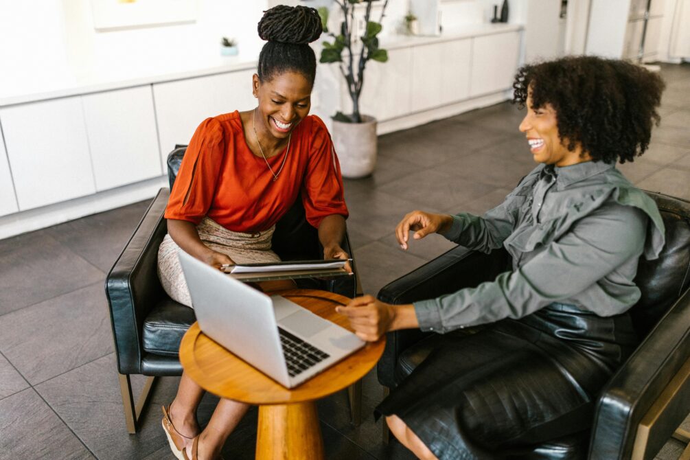 Duas mulheres trabalham juntas de forma descontraída em ambiente moderno e aconchegante, compartilhando ideias enquanto interagem com laptop e tablet em uma mesa redonda.